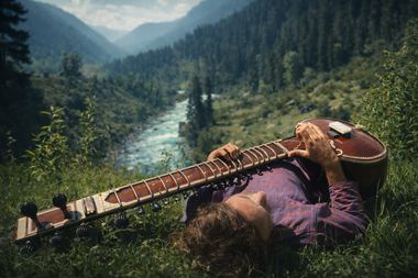 Yoav Fekete (YoYo Sitar) lying in a meadow playing his sitar above a Kashmir river valley