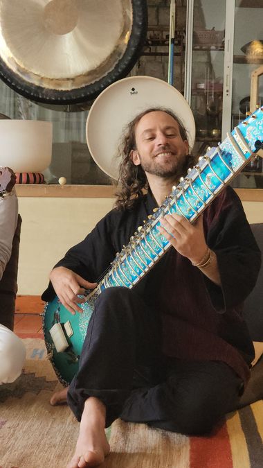 Yoav Fekete (YoYo Sitar) holding his blue sitar in his music room, beside a large gong