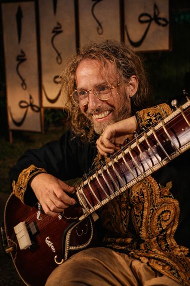 Yoav Fekete (YoYo Sitar) smiling with his ornate sitar before calligraphy panels at a Sufi festival